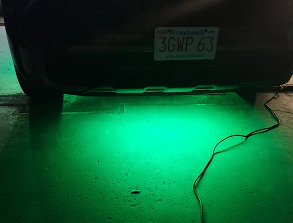 The underside of a car with a Massachusetts license plate emits a bright green glow on the floor, creating a sci-fi atmosphere. A cable is seen trailing off.