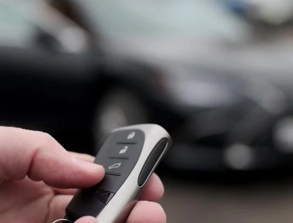 A hand holding a car key fob with a nearby blurred, dark-colored car. The focus is on the key fob, suggesting themes of convenience and technology.