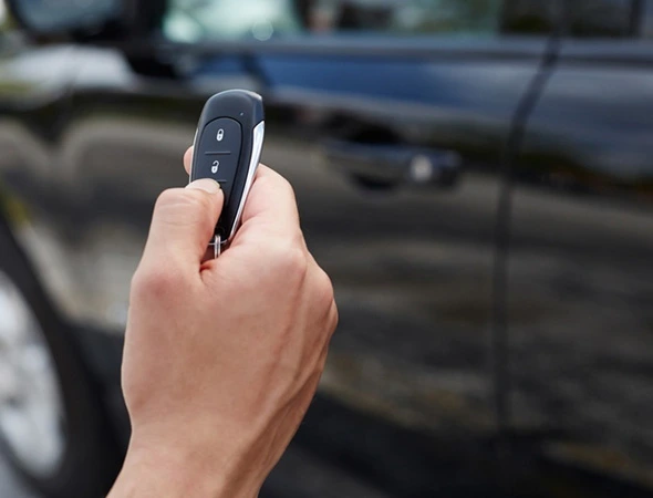 A hand holds a car key fob, pressing a button to lock or unlock a car in the background. The scene suggests convenience and technology.