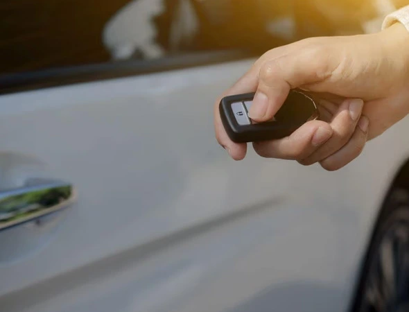 A hand holds a car key fob, pointing at a white car door under sunlight. The photo conveys a sense of modern convenience and security.