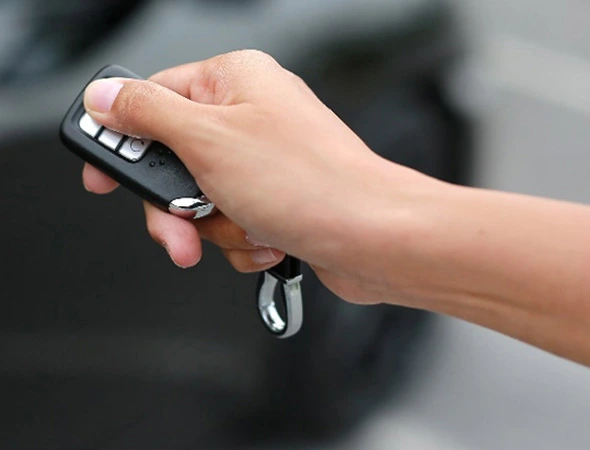 A hand holding a black car key fob, pressing a button. The background shows a blurred vehicle, suggesting unlocking a car with ease and convenience.
