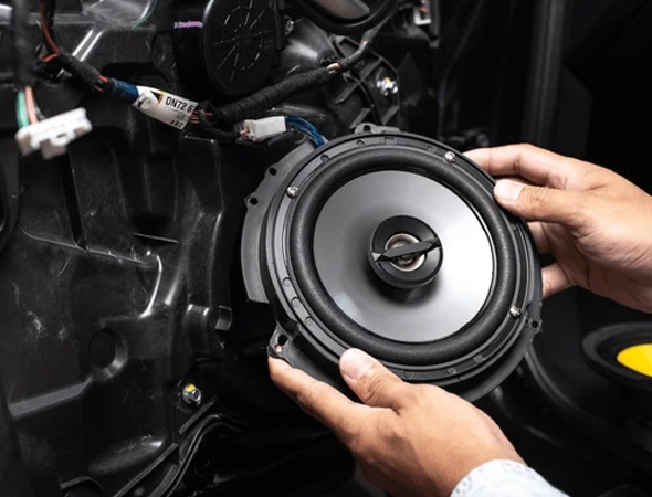 Installing a round car speaker into a vehicle interior, surrounded by wires and machinery. The focus is on the speaker, conveying precision.