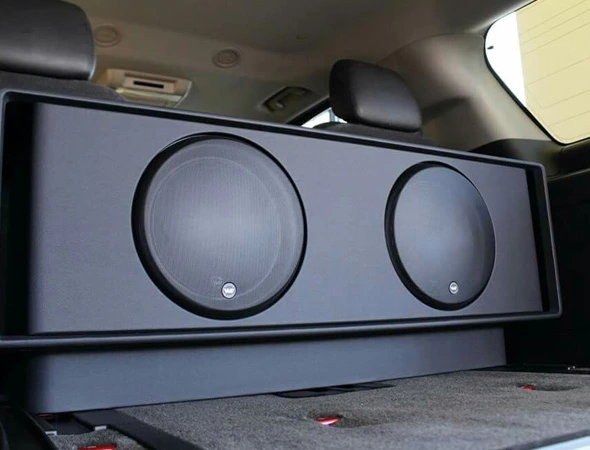 A large black speaker box with two circular subwoofers is placed in the back of a car. The interior is sleek, conveying a modern and powerful audio setup.