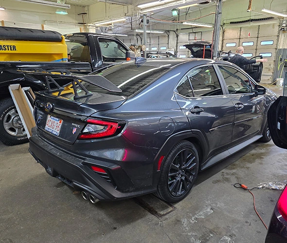 A sleek gray sedan with a large rear spoiler is parked inside a busy auto workshop. The setting is industrial, with various tools and vehicles around.