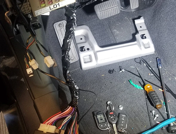 Car interior floor showing a car security system upgrade in progress, with exposed wiring harnesses, connectors, tools, and key fobs scattered on the carpet near the pedals.