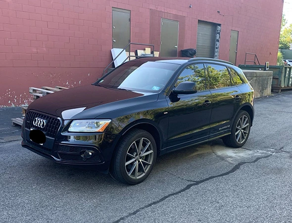 A black Audi SUV with tint window is parked on a paved lot beside a red brick building with utility doors. The scene is calm, with light reflecting off the car's surface.