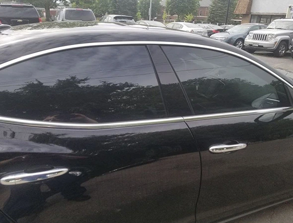 A black car with tinted windows and silver trim is parked in a lot. Reflections of trees and a cloudy sky are visible on the windows and body.